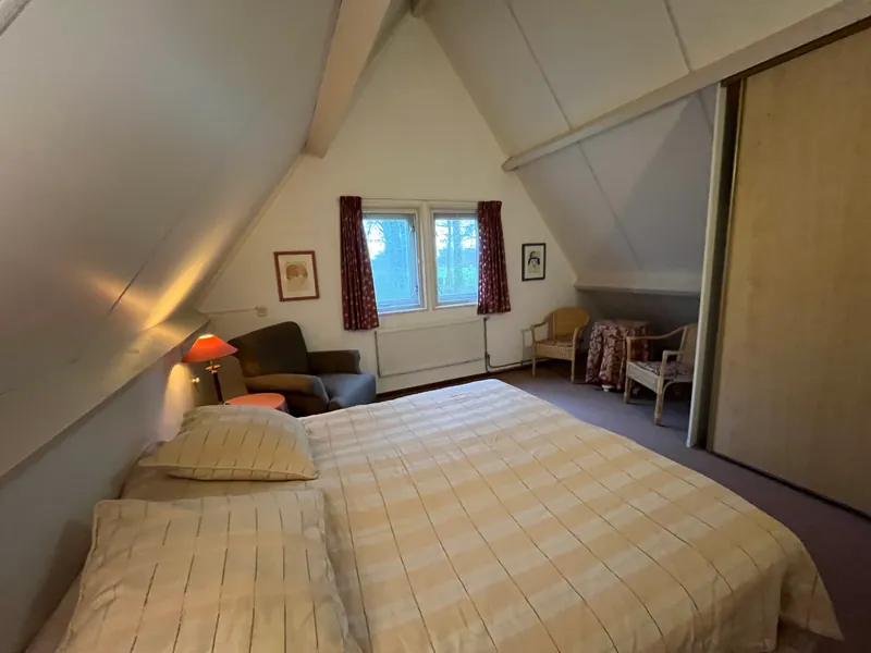 Boven, onder het schuine dak: de grote slaapkamer met twee ramen in de gevel, een breed bed en een leunstoel in de hoek.